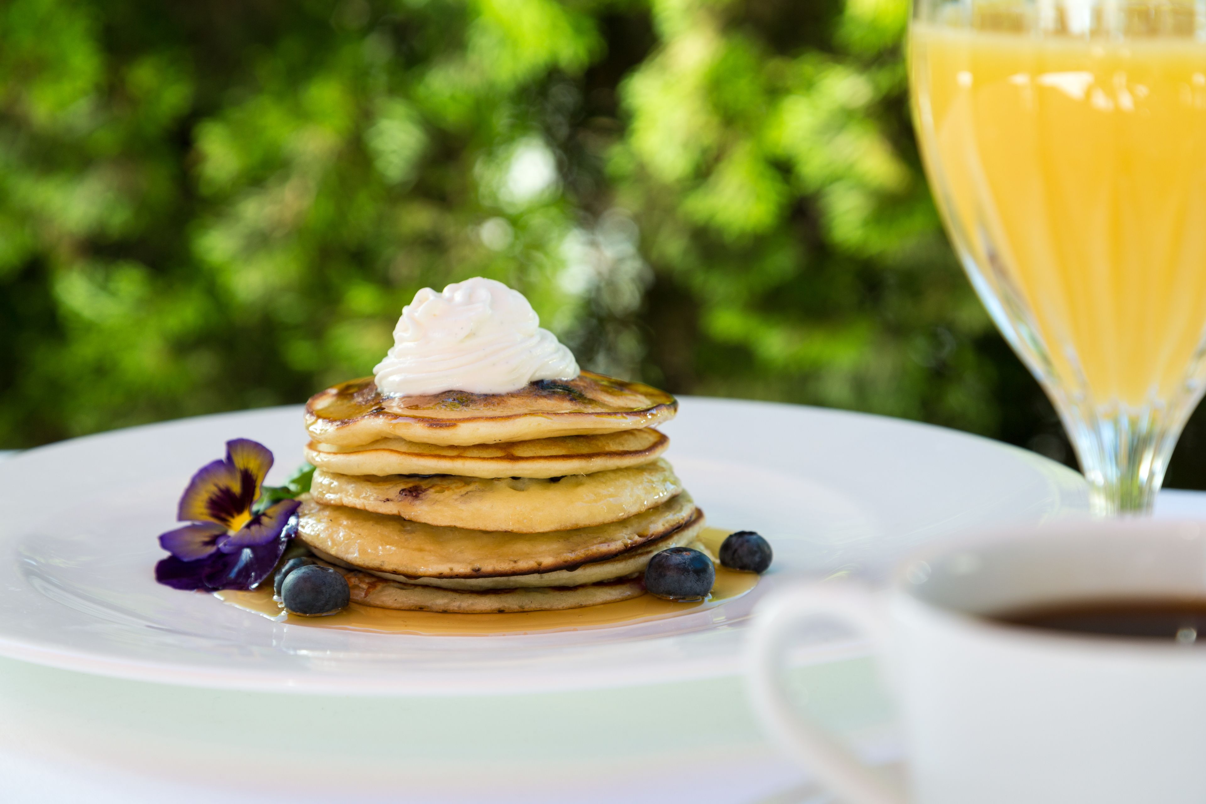 Una pila de tortitas con almíbar y arándanos sobre un plato blanco, con un vaso de jugo de naranja y una taza de café al fondo.