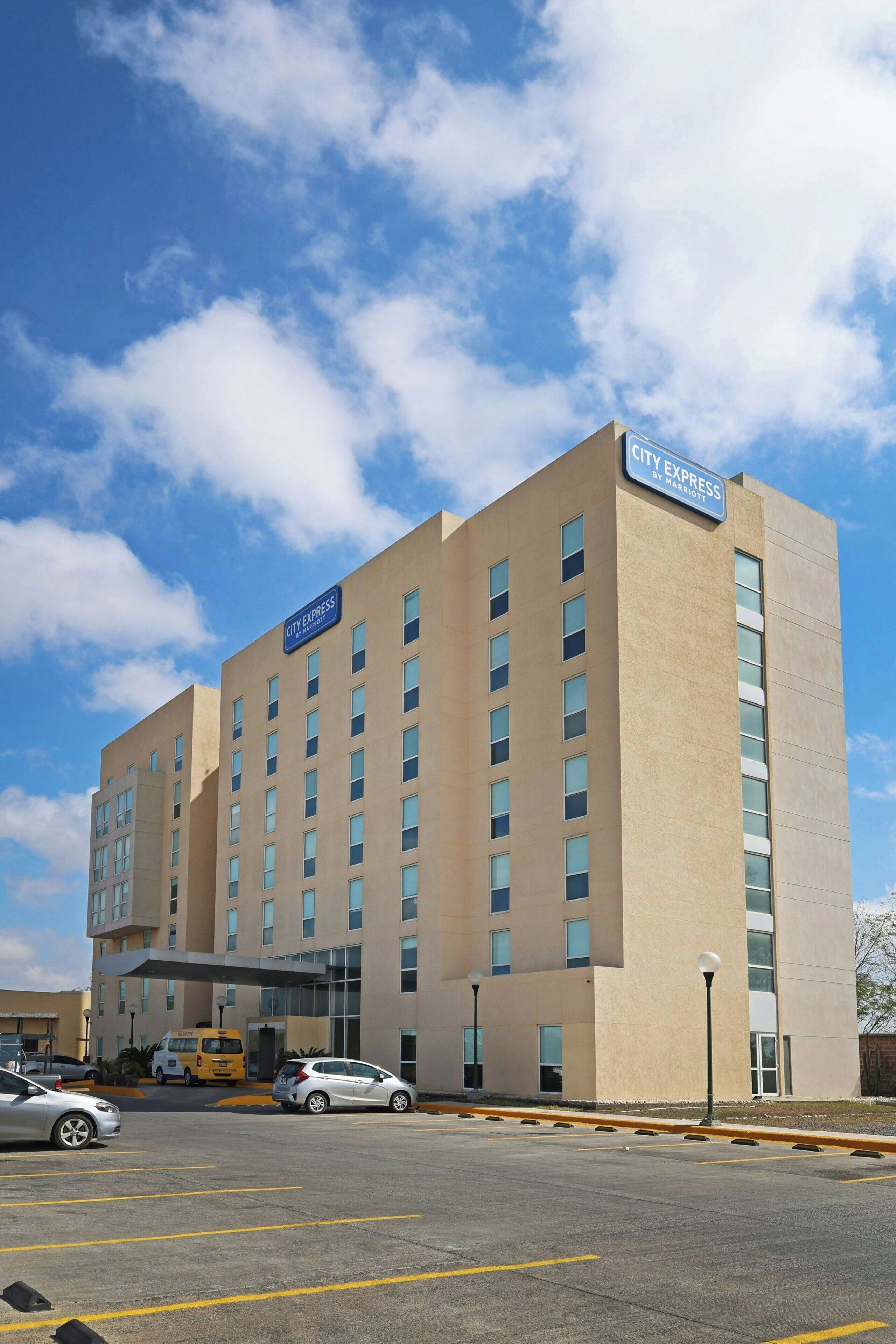A City Express hotel with a parking lot, a yellow shuttle, and a clear blue sky.