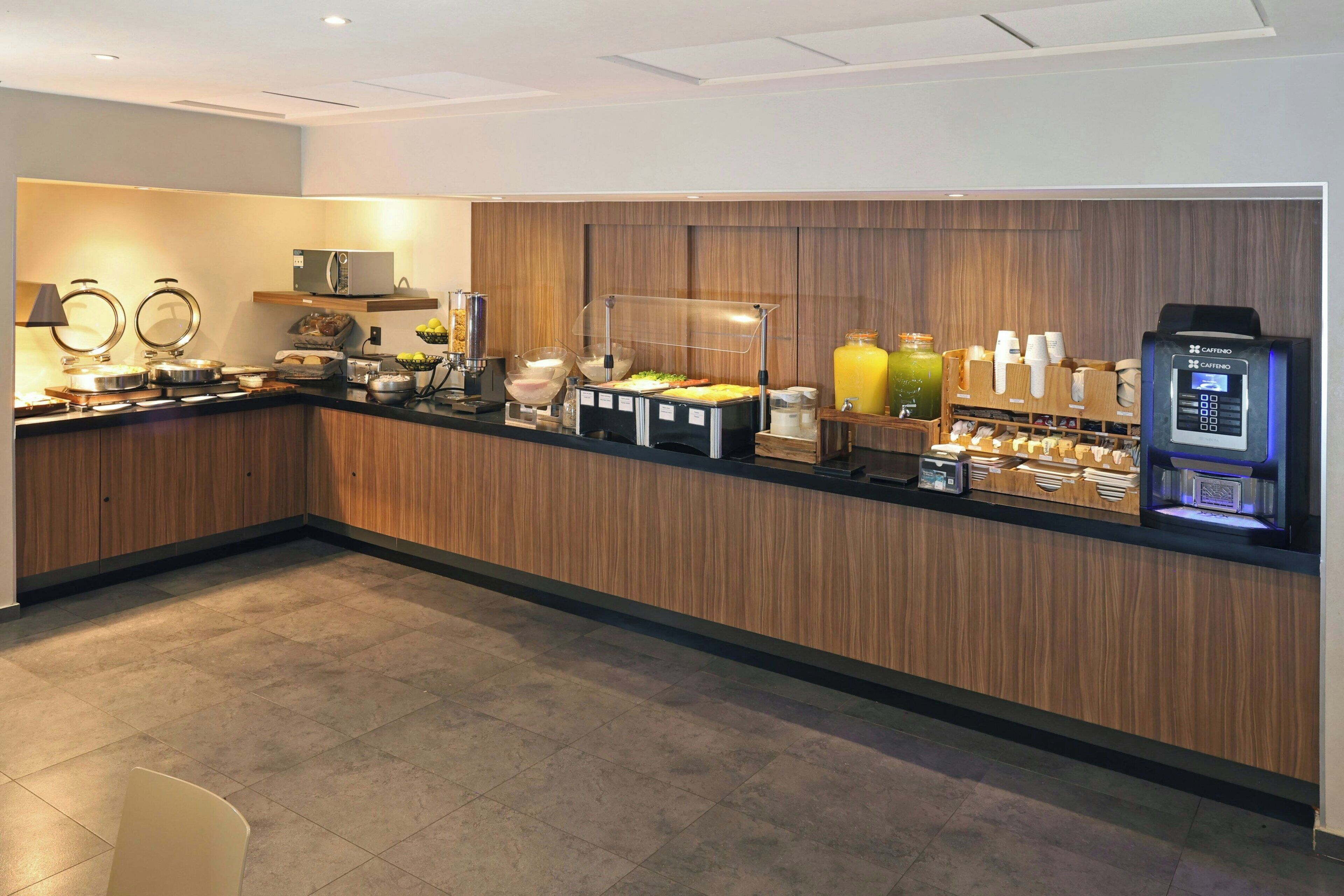 A modern breakfast bar with a coffee machine, fruit display, and a self-service beverage dispenser.