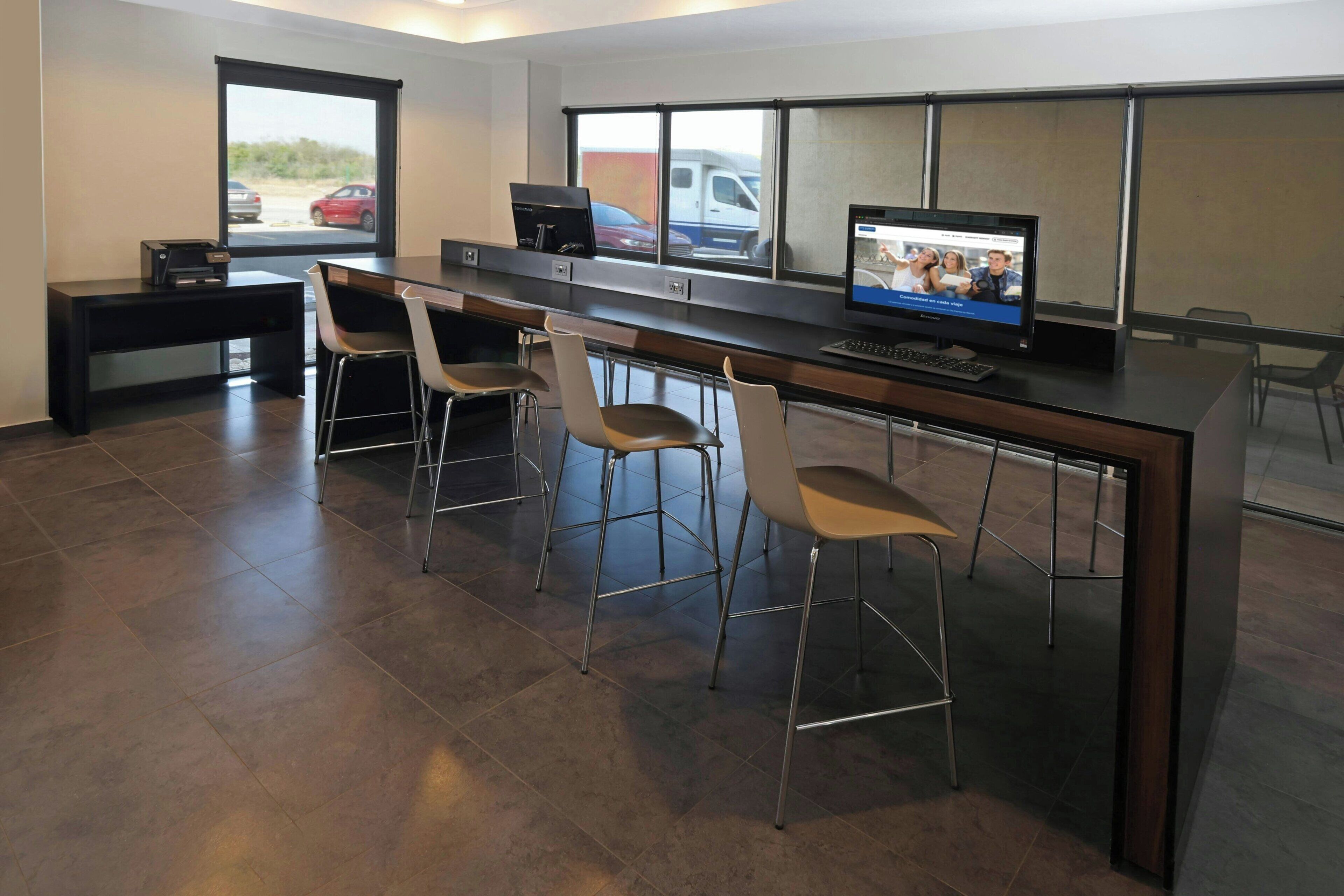 Un espacio de oficina moderno con un escritorio largo, tres sillas, una computadora y una ventana con vista a coches estacionados.