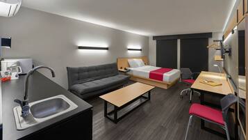 Une chambre d’hôtel moderne équipée d’une kitchenette, d’un canapé, d’un lit avec une couverture rouge, d’un bureau avec une chaise et d’une petite table.