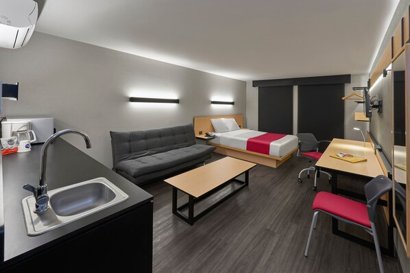 A modern hotel room with a kitchenette, a sofa, a bed with a red blanket, a desk with a chair, and a small table. - City Express by Marriott Reynosa Aeropuerto (Reynosa)