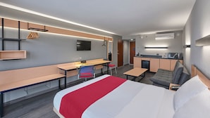 Ein modernes Hotelzimmer mit einem großen Bett, einem Schreibtisch, einem Sessel, einer Bank, einem kleinen Tisch, einem Fernseher und einer Küchenzeile.