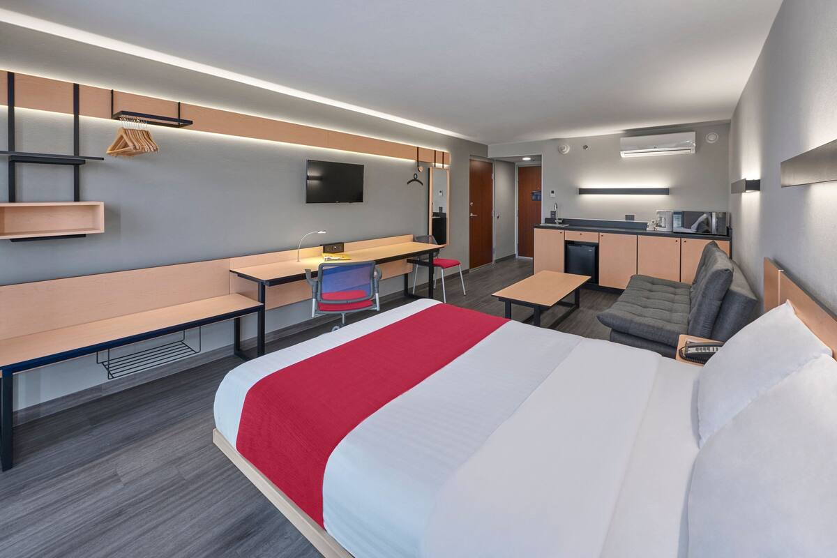 Ein modernes Hotelzimmer mit einem großen Bett, einem Schreibtisch, einem Sessel, einer Bank, einem kleinen Tisch, einem Fernseher und einer Küchenzeile.
