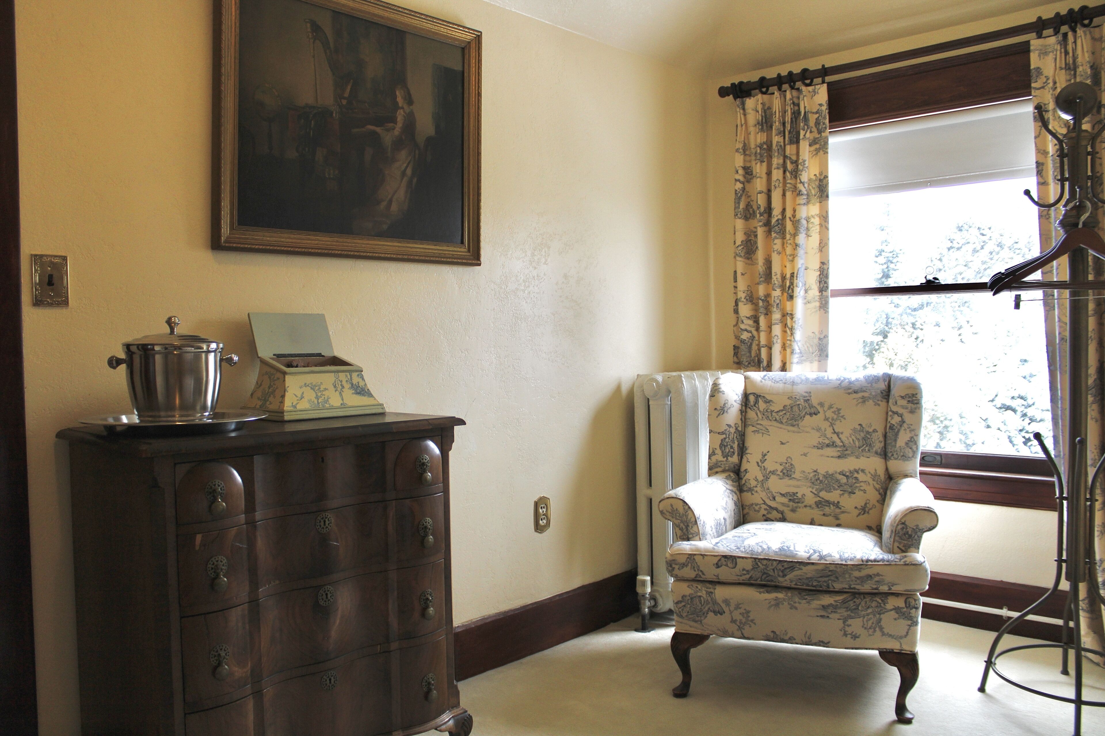 A room with a wooden dresser, a chair, a radiator, and a window with curtains.