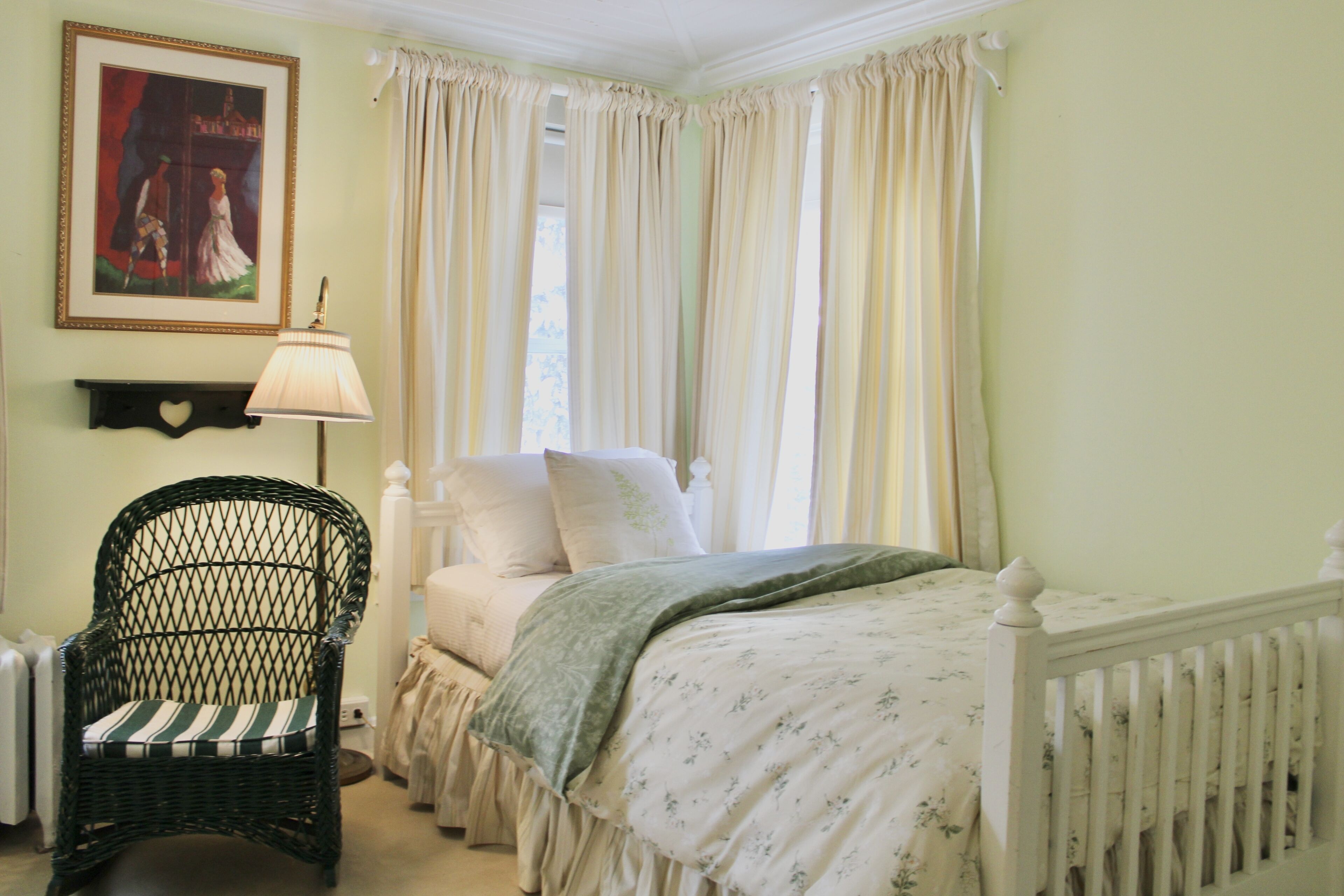 A bedroom with a bed, a wicker chair, a lamp, and a painting on the wall.