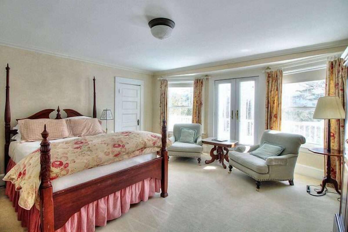 A bedroom with a four-poster bed, a chair, a small table, and a window with curtains.