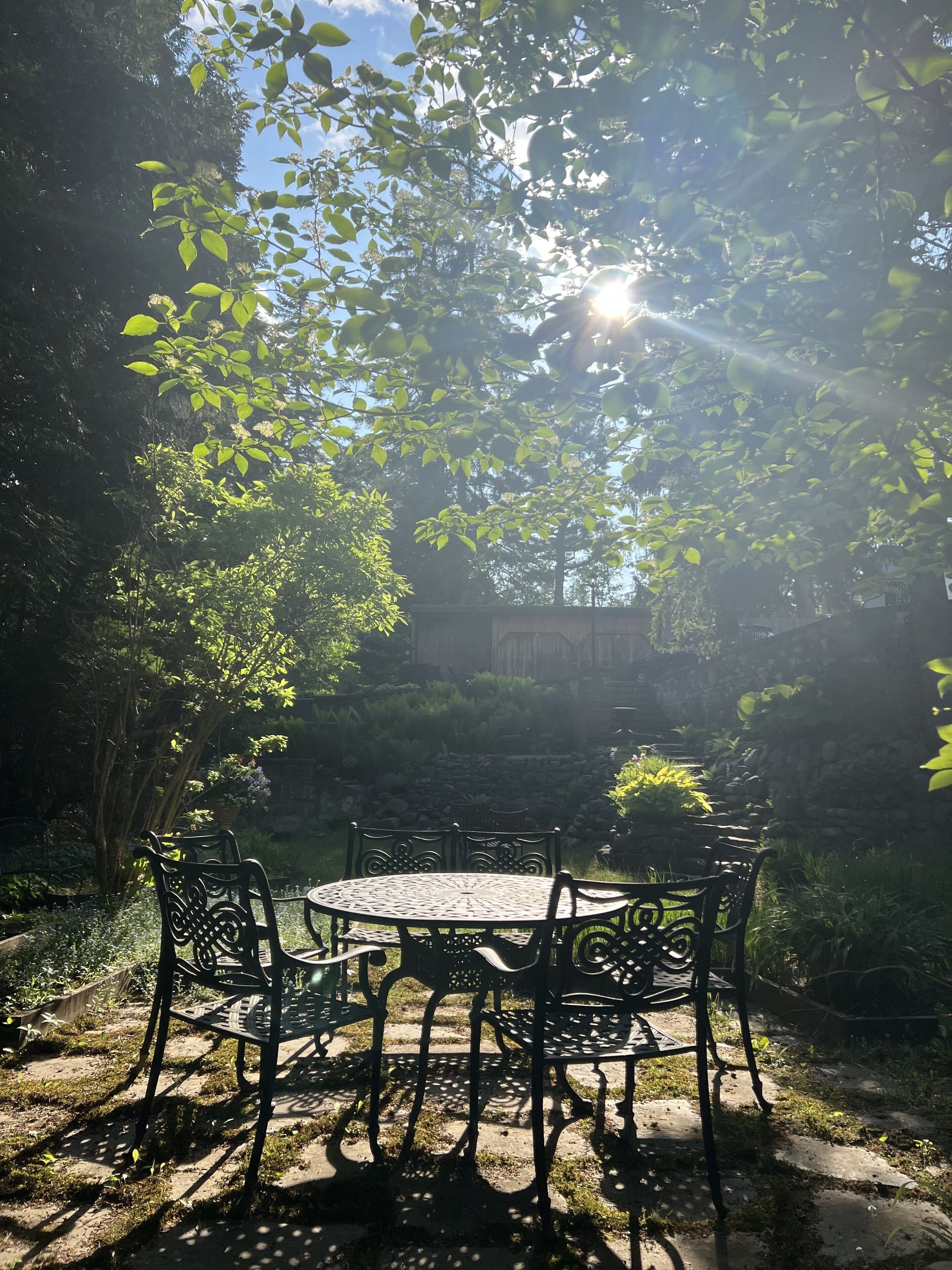 A garden patio with a round table and four chairs, surrounded by lush greenery and a stone wall.