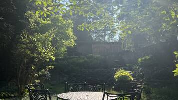 A garden patio with a round table and four chairs, surrounded by lush greenery and a stone wall.