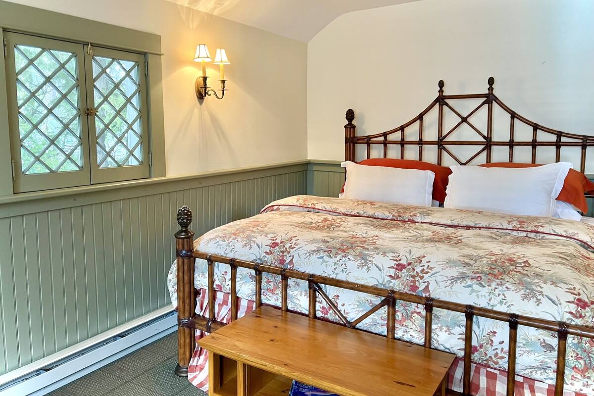 A bedroom with a wooden bed, floral bedding, a nightstand, a window with a view, and wall-mounted lamps.