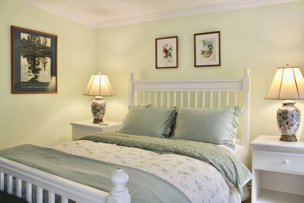 A bedroom with a white bed, green bedding, two bedside tables with lamps, and framed pictures on the wall.