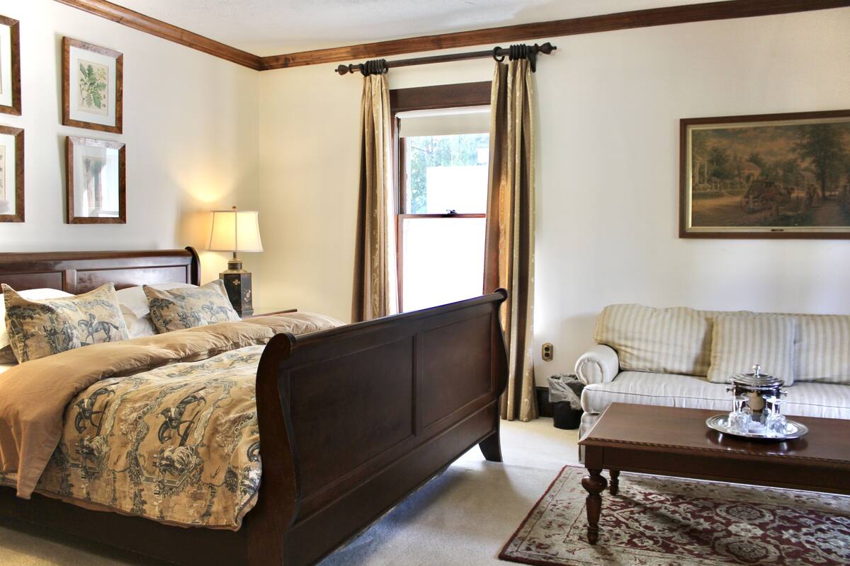 A bedroom with a bed, a wooden headboard, a nightstand with a lamp, a window with curtains, a sofa, and a coffee table.