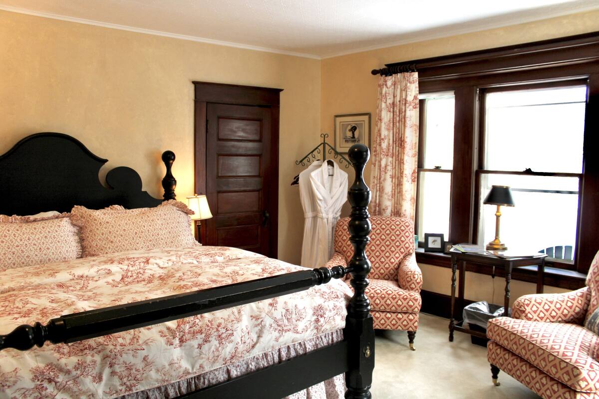 A bedroom with a four-poster bed, a sitting area with a chair and a table, and a window with curtains.