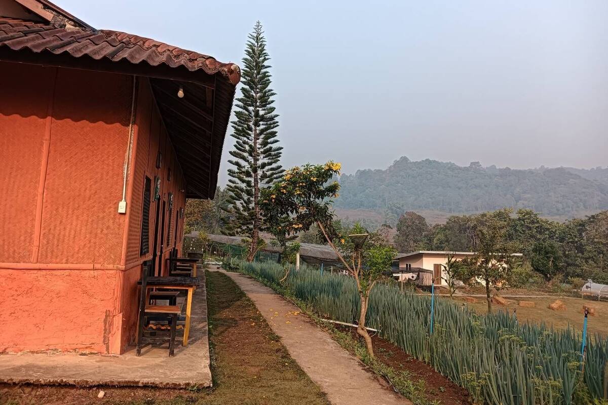 A red building with a tiled roof, a pathway leading to a garden, and a view of a hilly landscape in the background.