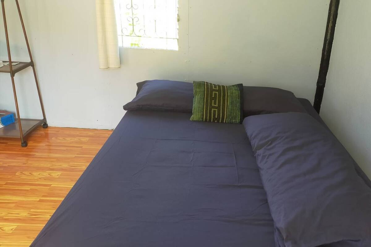 A single bed with a dark blue mattress and pillows, a window with metal bars, and a wooden floor.