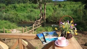Un banco de madera con una taza rosa y una cesta de flores, con vista a un río y un puente de madera en un paisaje verde exuberante.