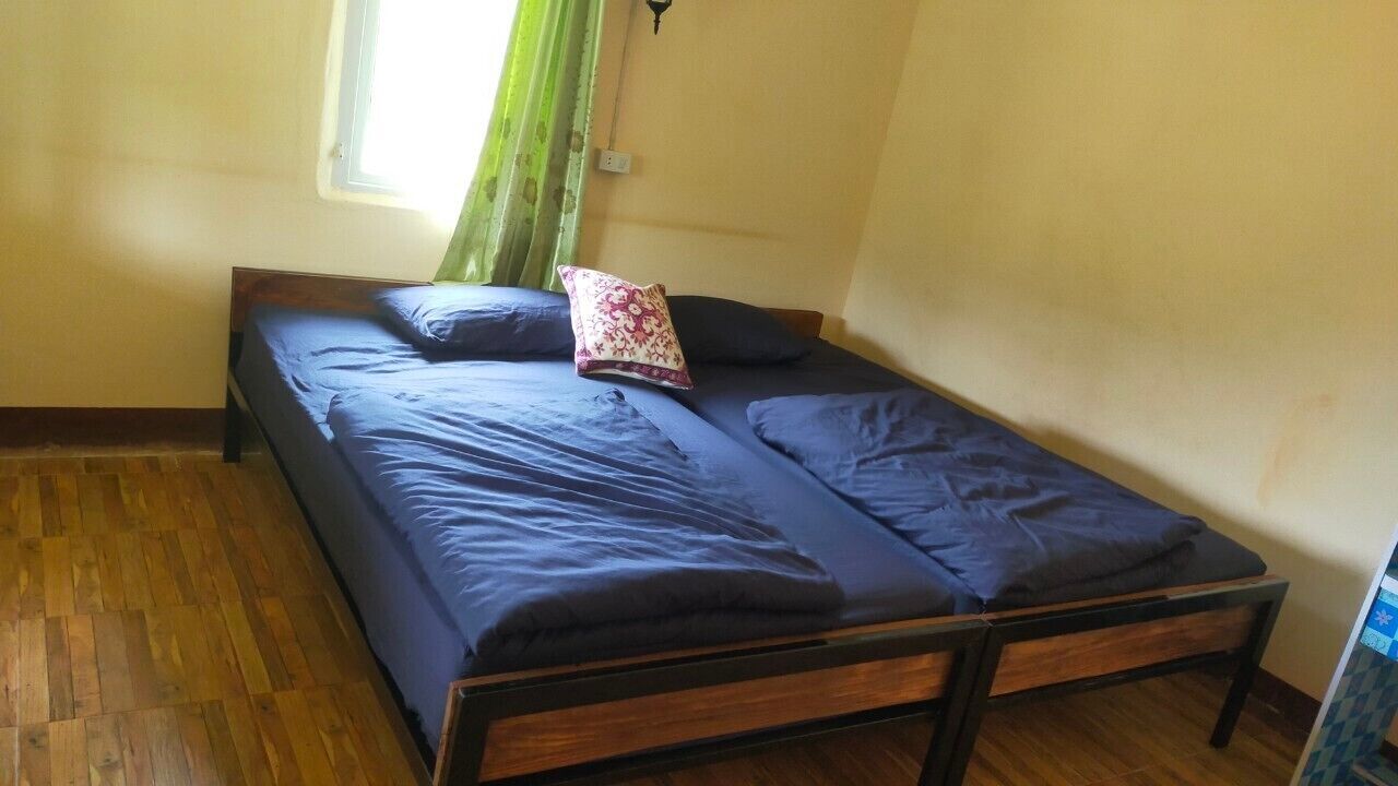Una cama individual con estructura de madera y ropa de cama oscura en una habitación con suelo de madera y una ventana con cortinas verdes.