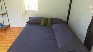 Una cama individual con colchón y almohadas azul oscuro, y una almohada verde a rayas, junto a una ventana con cortinas blancas.