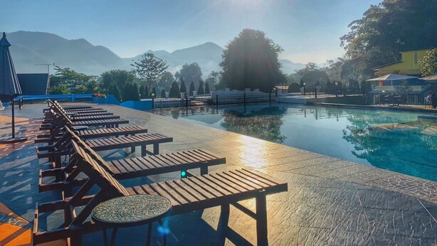 Un espace aménagé au bord de la piscine, avec des chaises longues en bois et une petite table, le tout entouré d’une clôture et d’arbres.