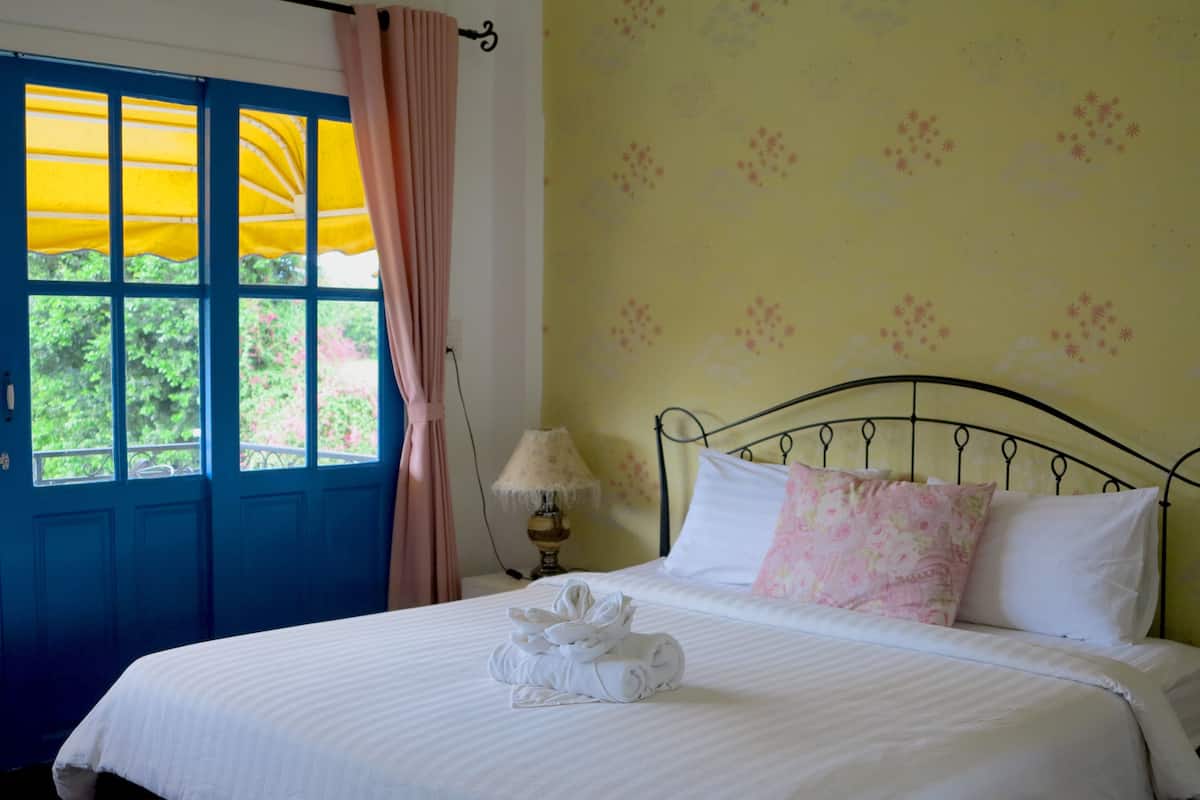 Ein Schlafzimmer mit Bett, einem Fenster mit Vorhängen, einer Lampe und blumigem Wandpapier.