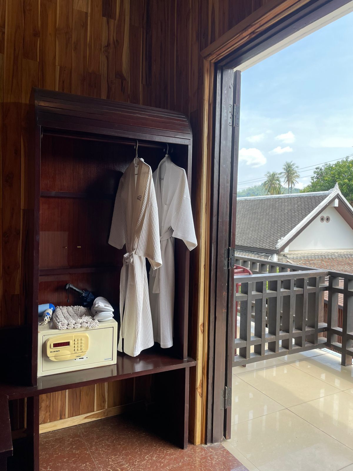 A wooden wardrobe with white robes hanging inside, a digital clock, and a view of a traditional house through an open door.