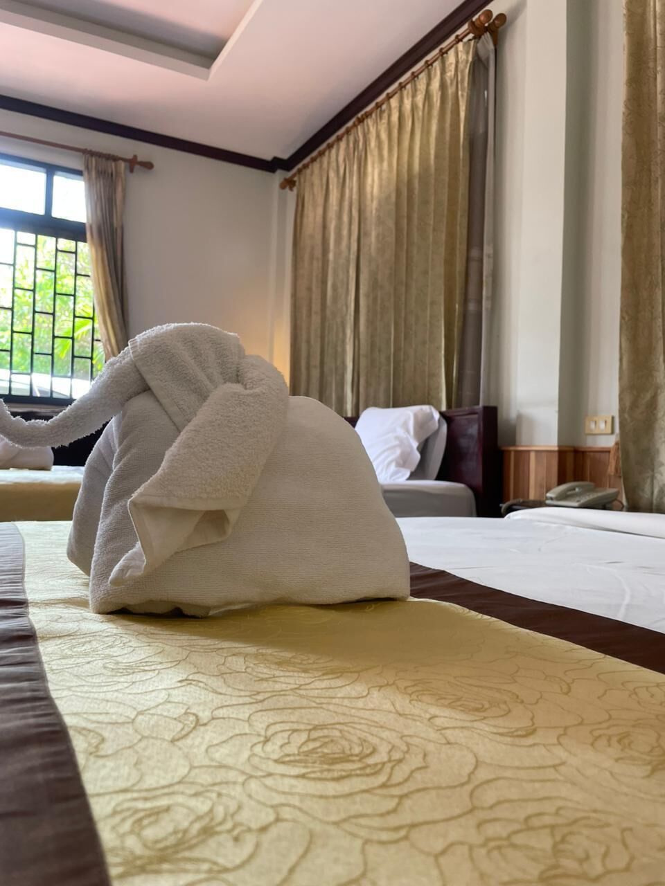 A hotel room with a neatly folded white towel on the bed, a window with curtains, and a bed with a patterned bedspread.