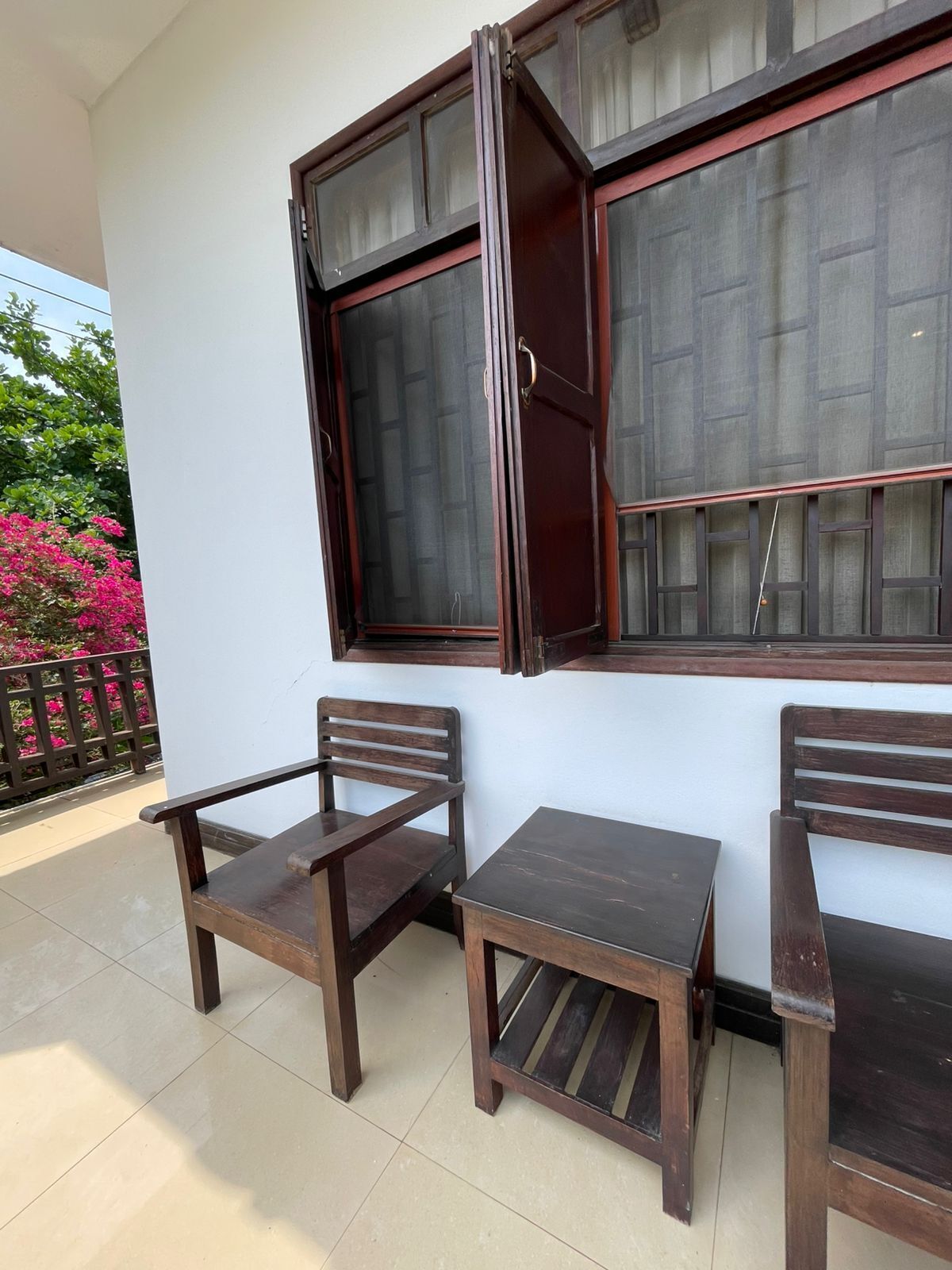 Een balkon met twee houten stoelen en een kleine tafel, een raam met houten kader, en uitzicht op roze bloemen en een hek.