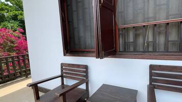 Een balkon met twee houten stoelen en een kleine tafel, een raam met houten kader, en uitzicht op roze bloemen en een hek.