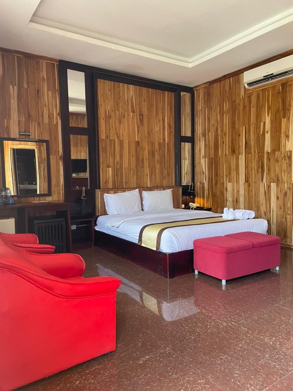 A hotel room with a wooden paneled wall, a bed with white linens, a red armchair, a red ottoman, and a desk with a mirror.