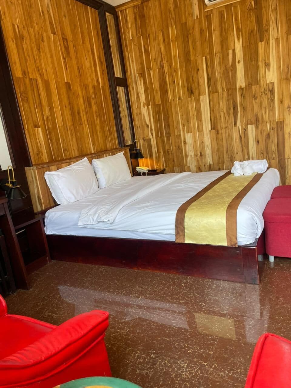 Een hotelkamer met een houten wand, een bed met witte lakens, een nachtkastje met een lamp en een rode stoel.
