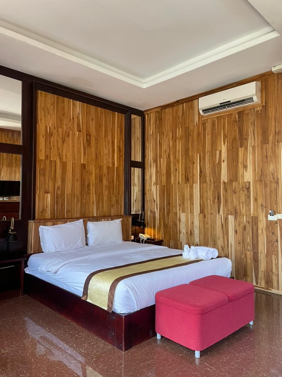 A hotel room with a wooden panel wall, a large bed with white bedding, a red ottoman, and a ceiling-mounted air conditioning unit.