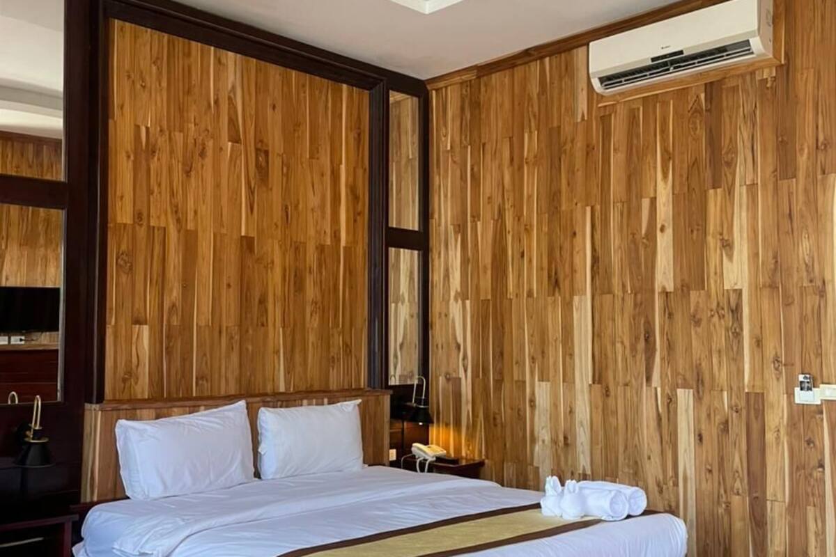 A hotel room with a wooden panel wall, a large bed with white bedding, a red ottoman, and a ceiling-mounted air conditioning unit.