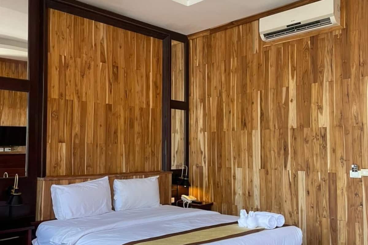 A hotel room with a wooden panel wall, a large bed with white bedding, a red ottoman, and a ceiling-mounted air conditioning unit.