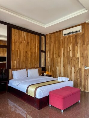 A hotel room with a wooden panel wall, a large bed with white bedding, a red ottoman, and a ceiling-mounted air conditioning unit. - Luang Prabang Inn (Luang Prabang)