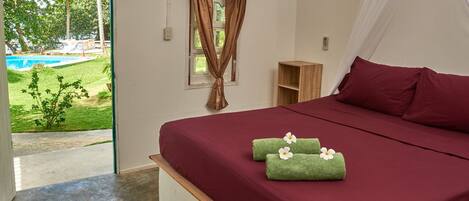 Un dormitorio con una cama grande, una mesita de noche de madera, un estante pequeño de madera y vistas a una piscina y vegetación.