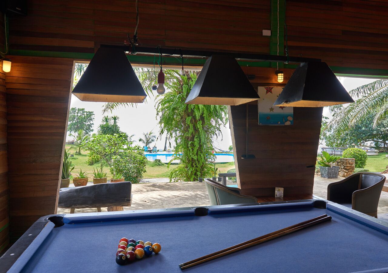 Una mesa de billar con bolas y tacos, paredes de madera, lámparas colgantes y vistas a una piscina y vegetación al exterior.
