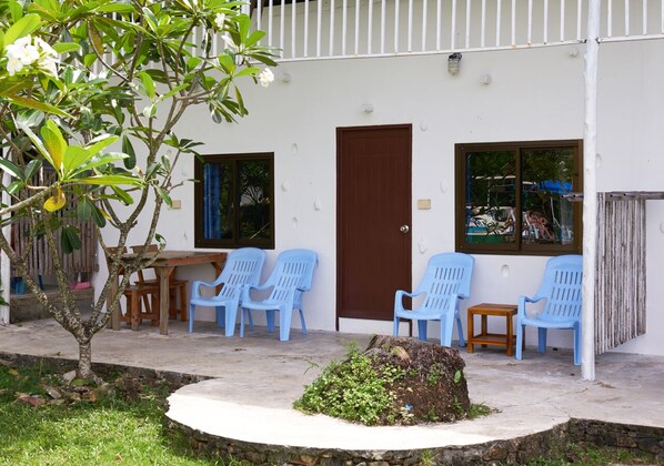 Un edificio pequeño con fachada blanca, una puerta marrón y un ventanal. Hay sillas de plástico azul y una mesa de madera afuera.