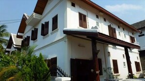 A two-story house with a white exterior, dark brown roof, and multiple windows with wooden shutters. - Mao Phashok Guesthouse (Luang Prabang)