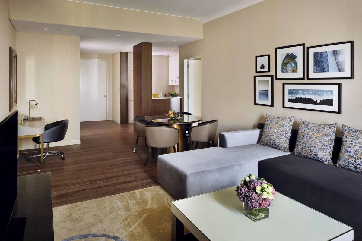 Una habitación de hotel moderna con un sofá, una mesa de comedor con sillas, un televisor y una mesa de centro con flores.