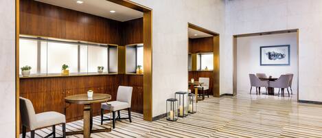 A modern hotel lobby with wooden paneling, a marble floor, and a seating area with tables and chairs.