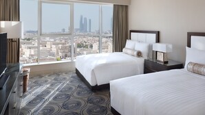 A hotel room with two beds, a large window with a city view, a flat-screen TV, and a minibar.