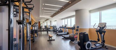 A modern gym with various exercise equipment, large windows, and a wooden floor.