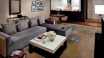 A modern hotel room with a grey sofa, a small round table with a vase of flowers, a coffee table, and a dining area with a bar.