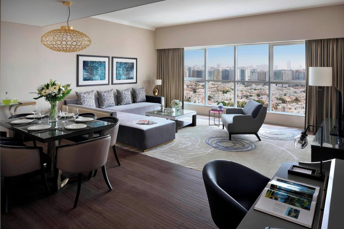 Habitación de hotel moderna con zona de comedor, sofá, silla y un amplio ventanal con vistas a la ciudad.