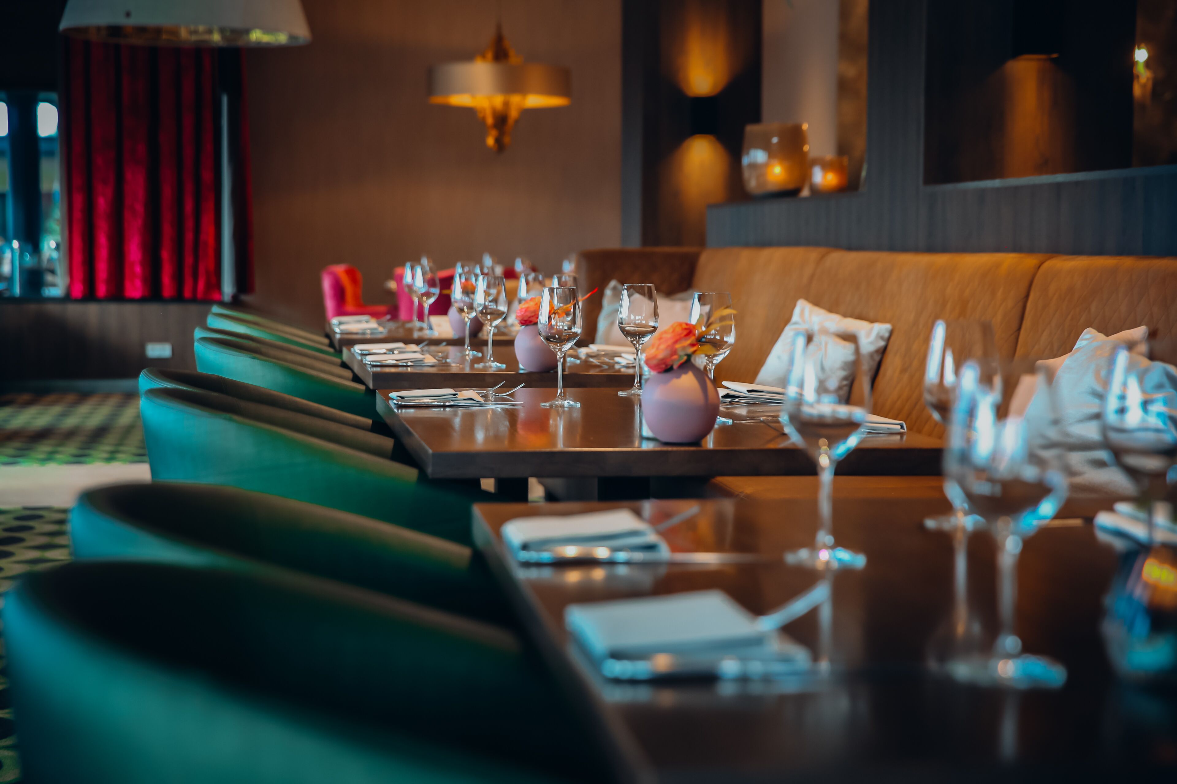 A restaurant with a long table set for dining, green upholstered seating, and a warm ambient lighting.