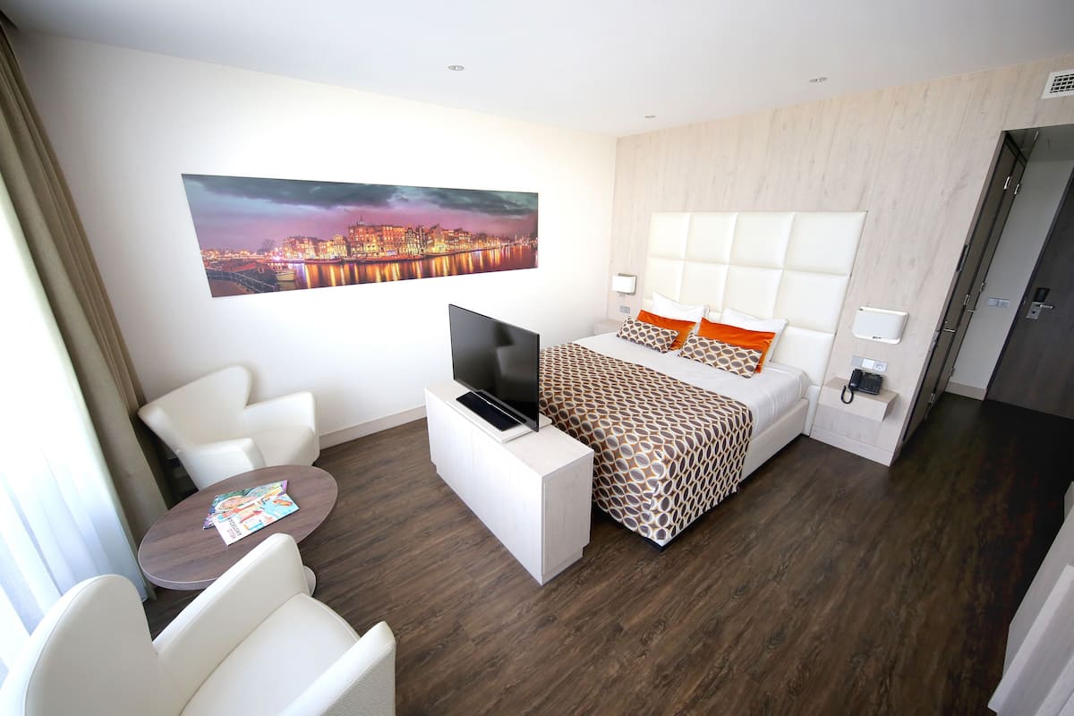 A modern hotel room with a large bed, a TV on a white console, a seating area with a table, and a cityscape painting on the wall.