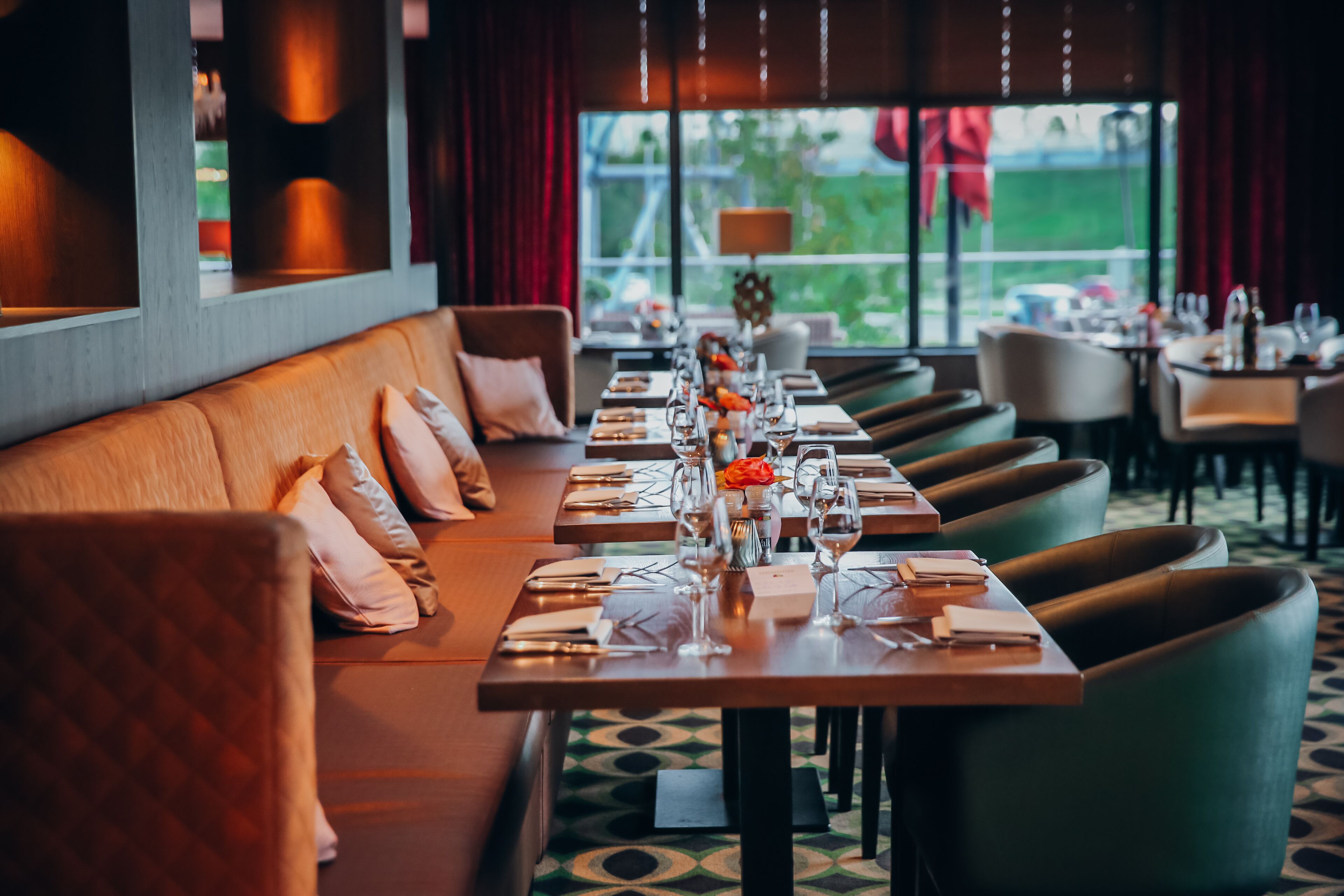 En restaurant med et langt spisebord, polstrede siddepladser og udsigt til det fri.