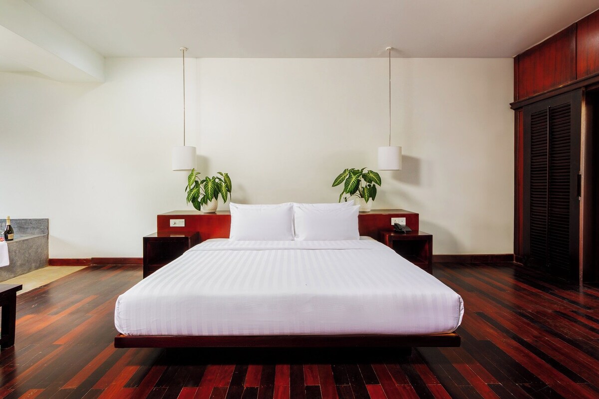 Une chambre d’hôtel avec un grand lit, deux tables de chevet avec des lampes, un plancher en bois et une plante sur chaque table.