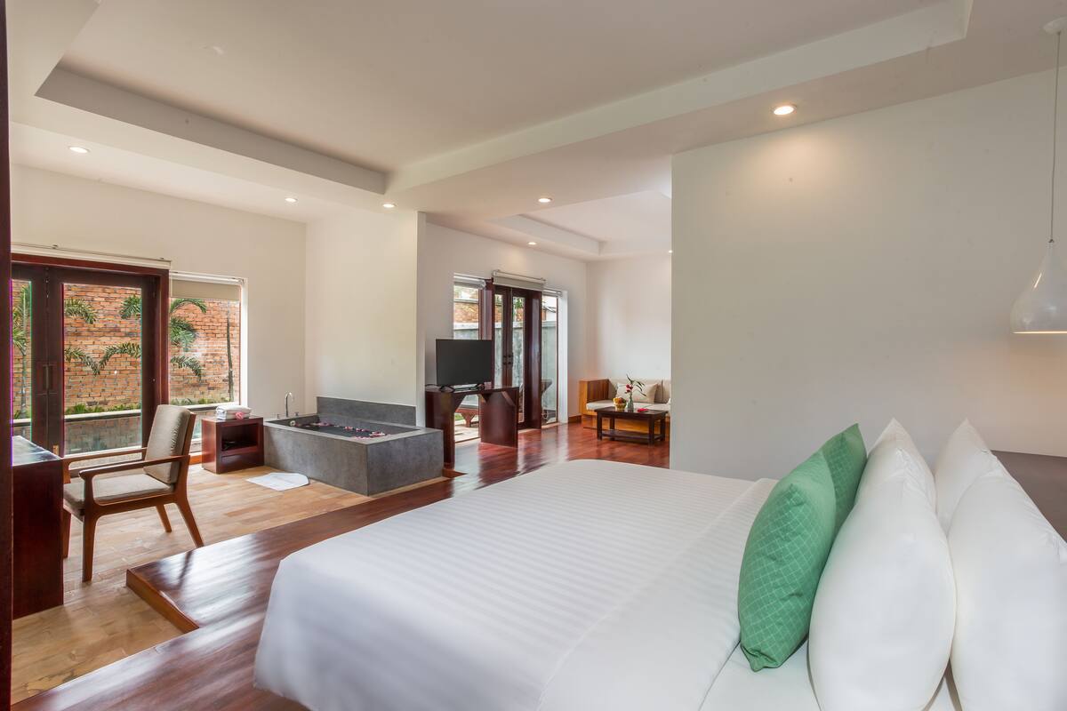 Une chambre d’hôtel spacieuse avec un grand lit, un coin salon comprenant un canapé et un fauteuil, une petite table et une vue sur un mur de briques et la verdure environnante.