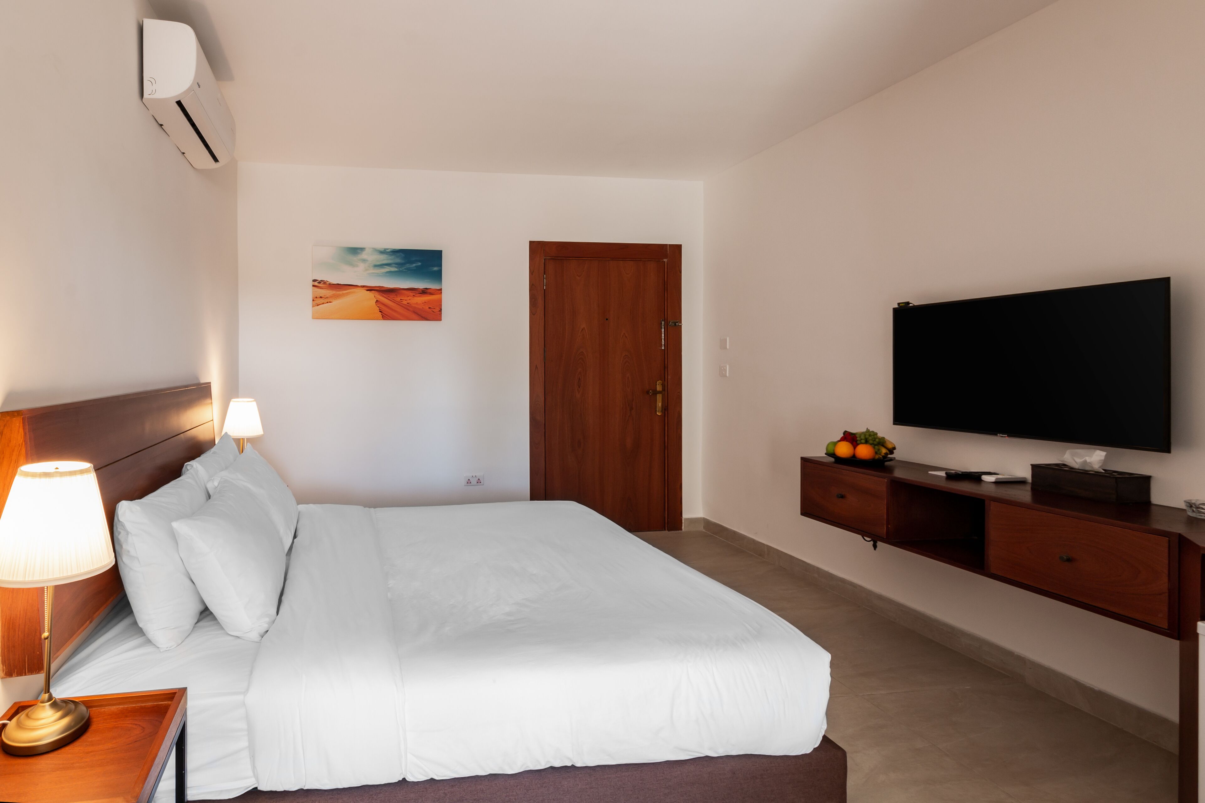 A hotel room with a bed, a TV, a wooden dresser, a lamp, and a painting on the wall.